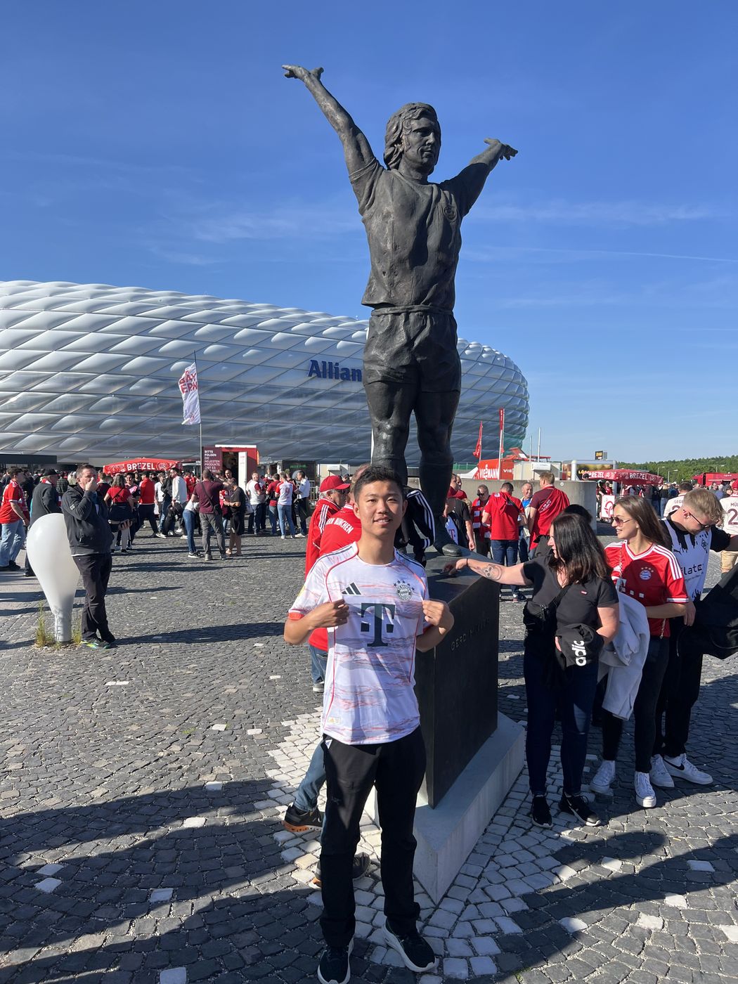 Thomas in a Bayern München jersey beside a statue outside the Allianz Arena, among crowds of fans