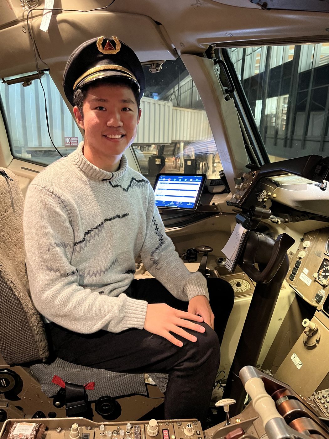 Thomas wearing a pilot’s cap, seated in an airliner cockpit at the gate